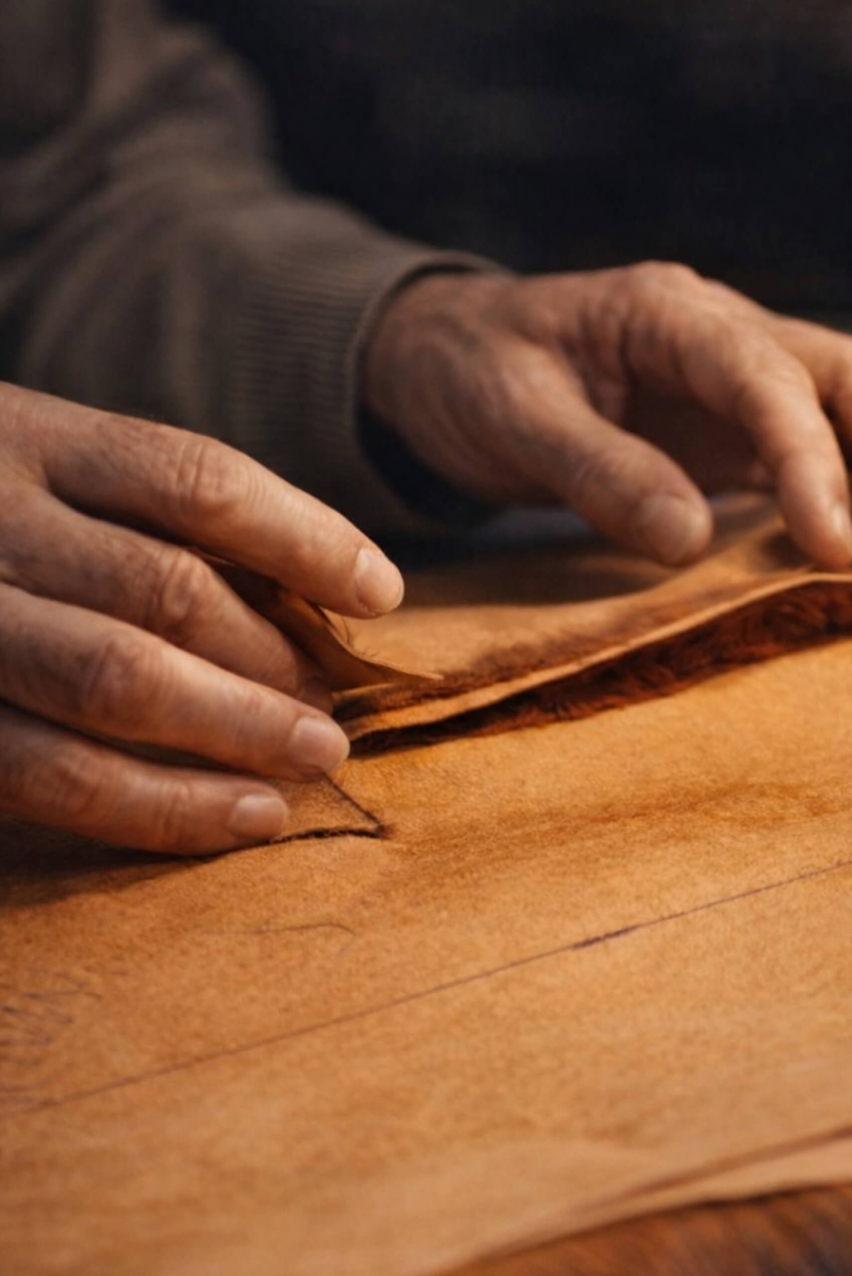 A close-up shot of an artisan's hands carefully handling and examining tan-colored raw leather pieces. The image highlights the natural texture of the leather and the meticulous detail of traditional hand-craftsmanship in a workshop setting.